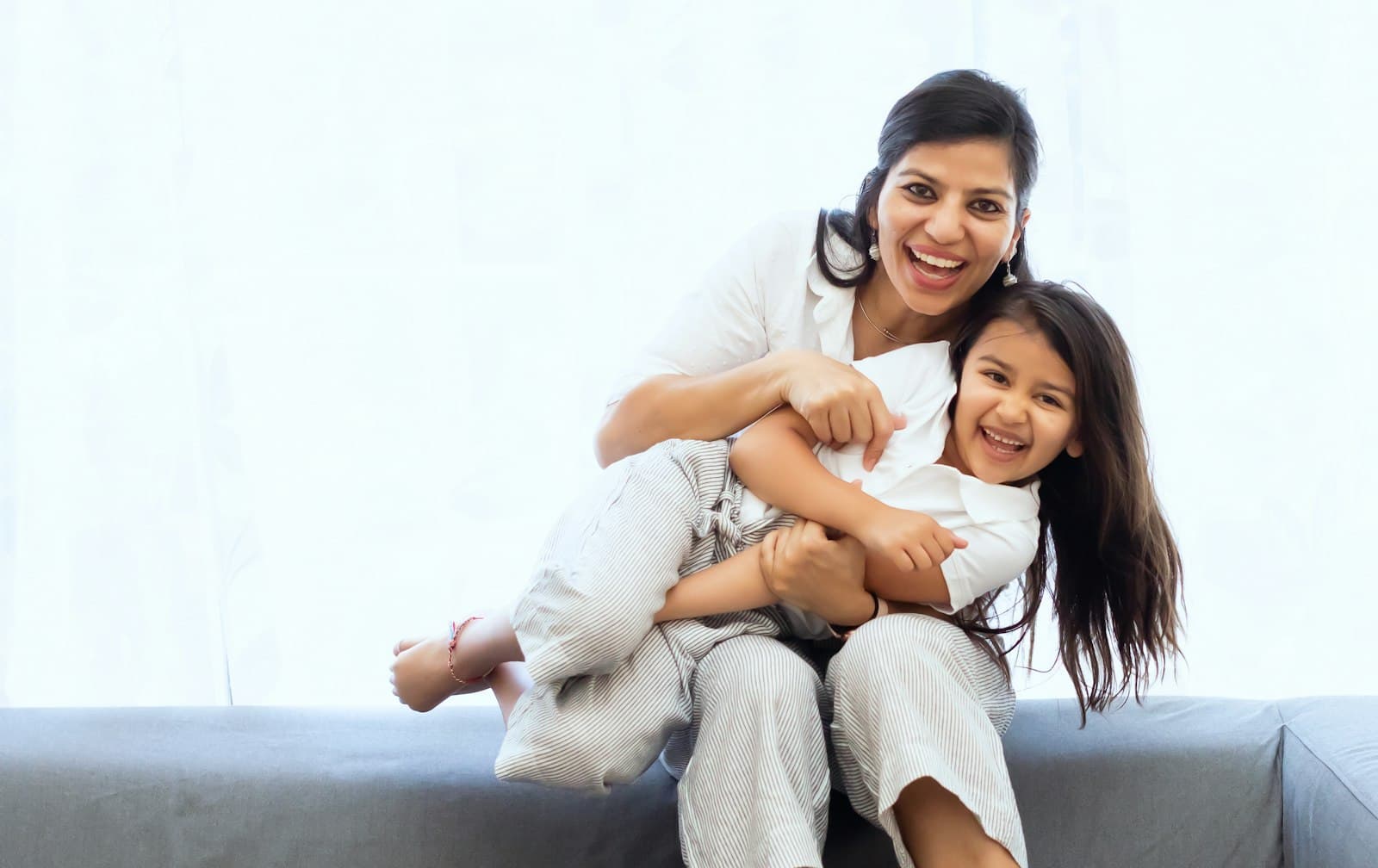 A mother holding her laughing daughter on a sofa at home.